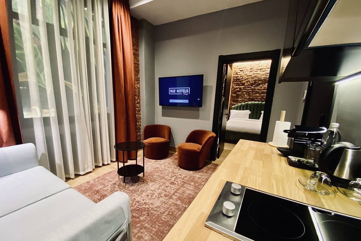 Quarto de hotel moderno com televisão de tela plana, sofá, duas poltronas, máquina de café e pequena cozinha.