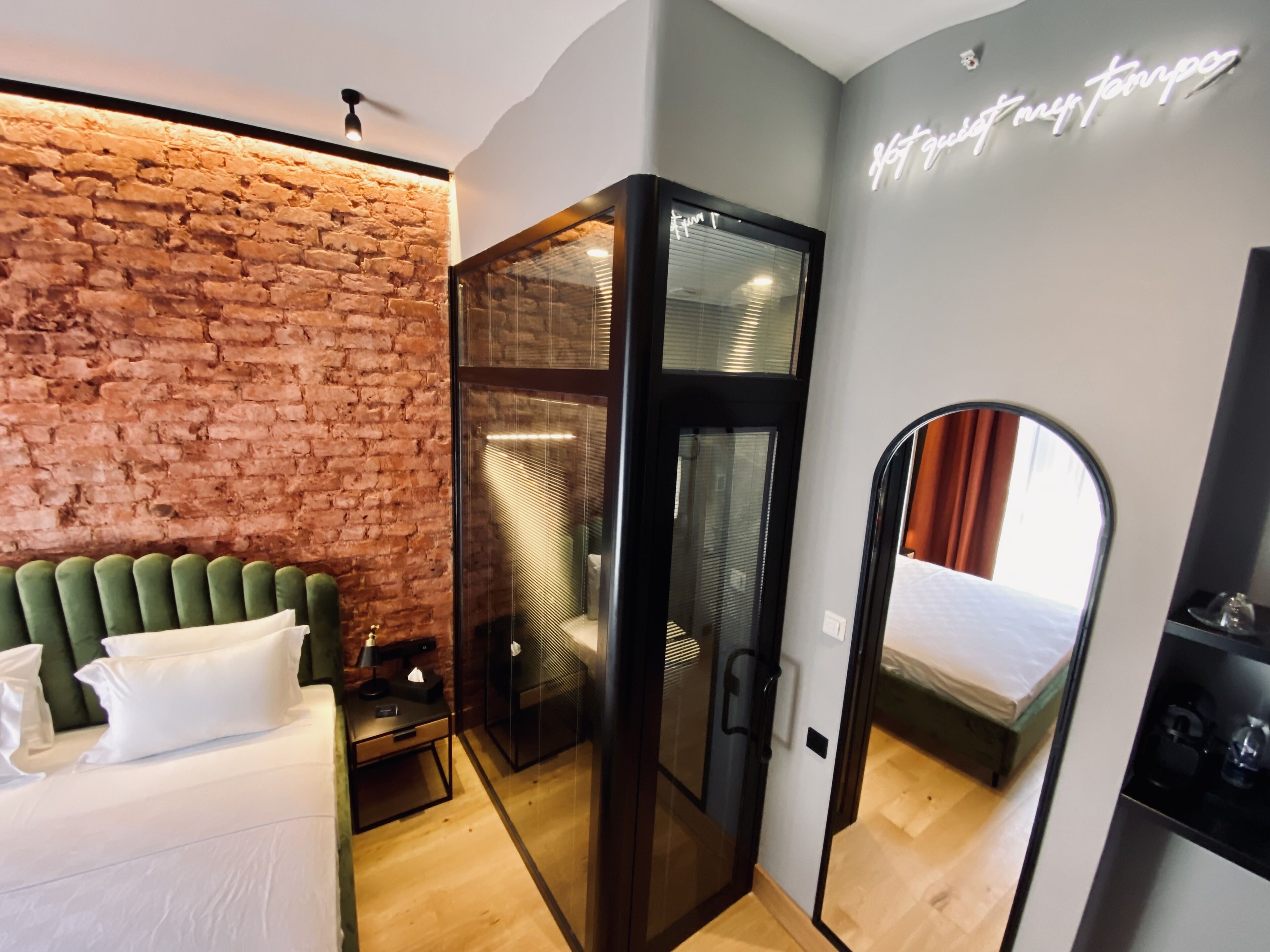 A modern hotel room with a brick wall, a green upholstered headboard, a glass partition, and a mirror reflecting the room.