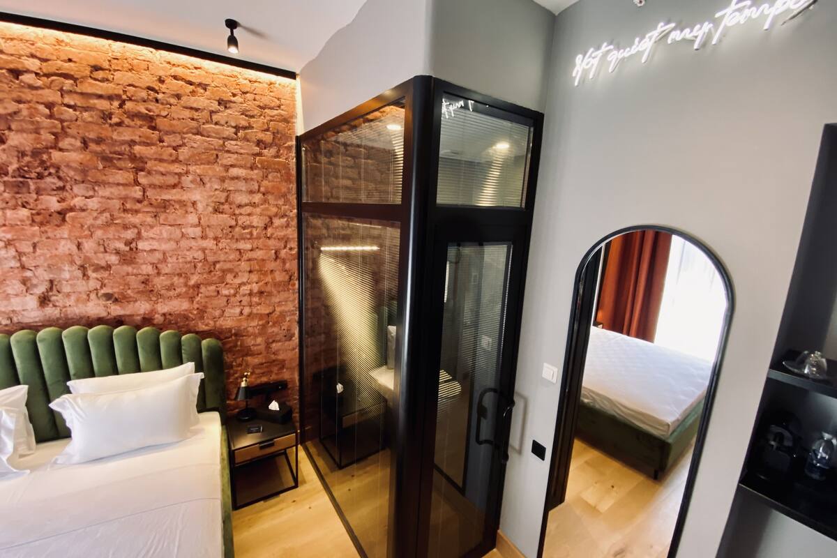 Quarto de hotel moderno com parede de tijolos, cabeceira estofada em verde, divisória de vidro e um espelho refletindo o aposento.
