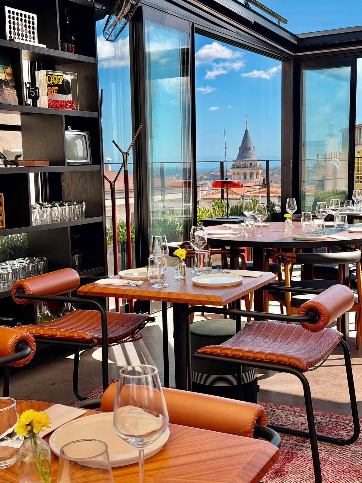 A restaurant with a view of a cityscape, featuring tables set for dining, leather chairs, and a large window.