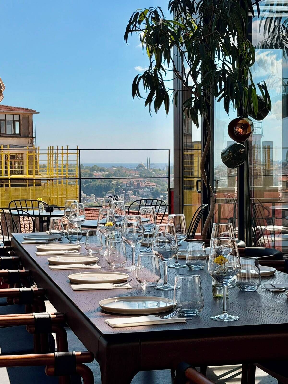 A dining table set for a meal with a city view, a plant, and a metal sculpture.