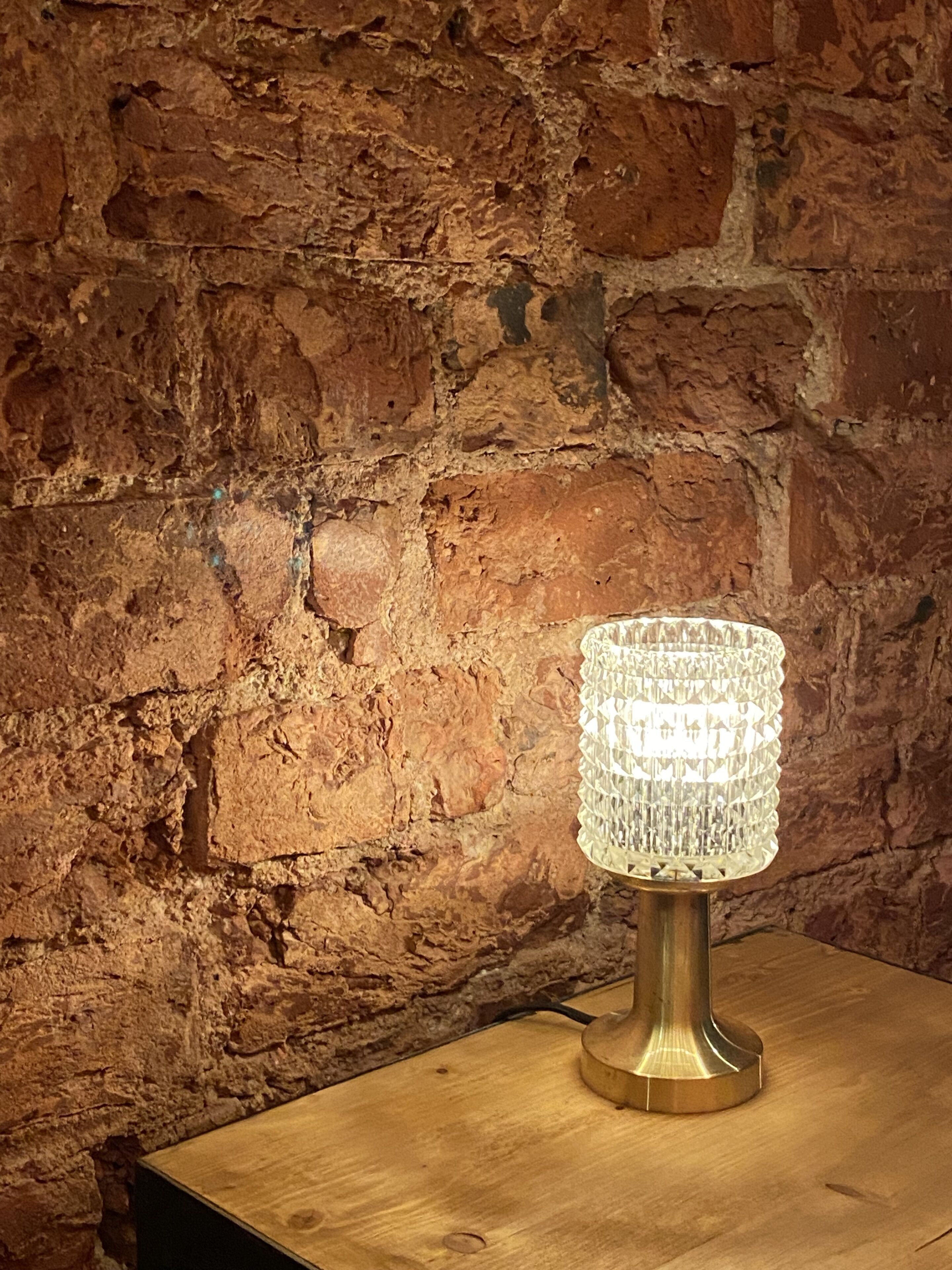 A table lamp with a crystal shade on a wooden table against a brick wall.