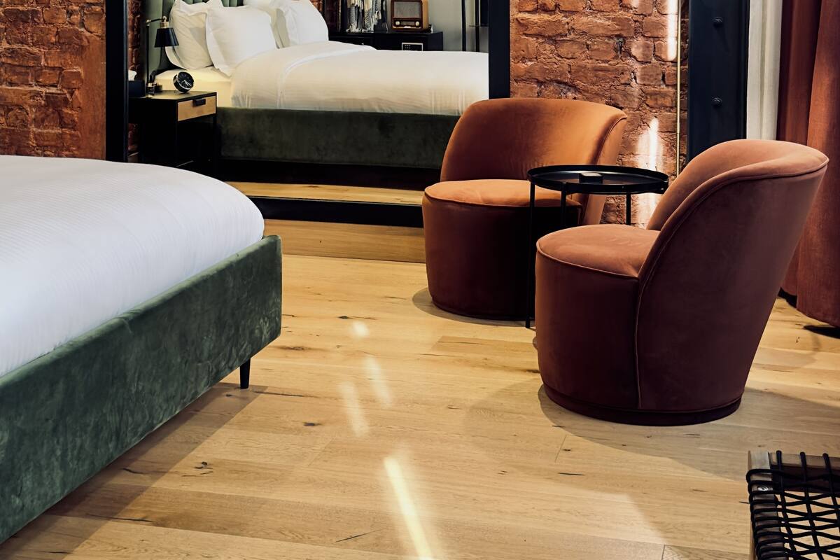 Quarto de hotel moderno com parede de tijolos, uma cama com cabeceira verde, uma mesa pequena, duas poltronas e uma mesa de cabeceira com abajur.
