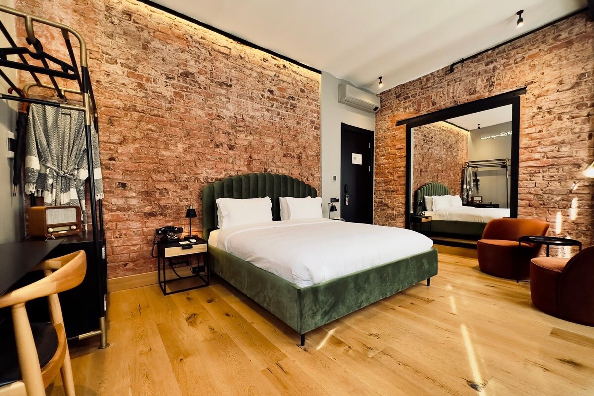 A modern hotel room with a brick wall, a large bed, a desk with a chair, and a mirror.