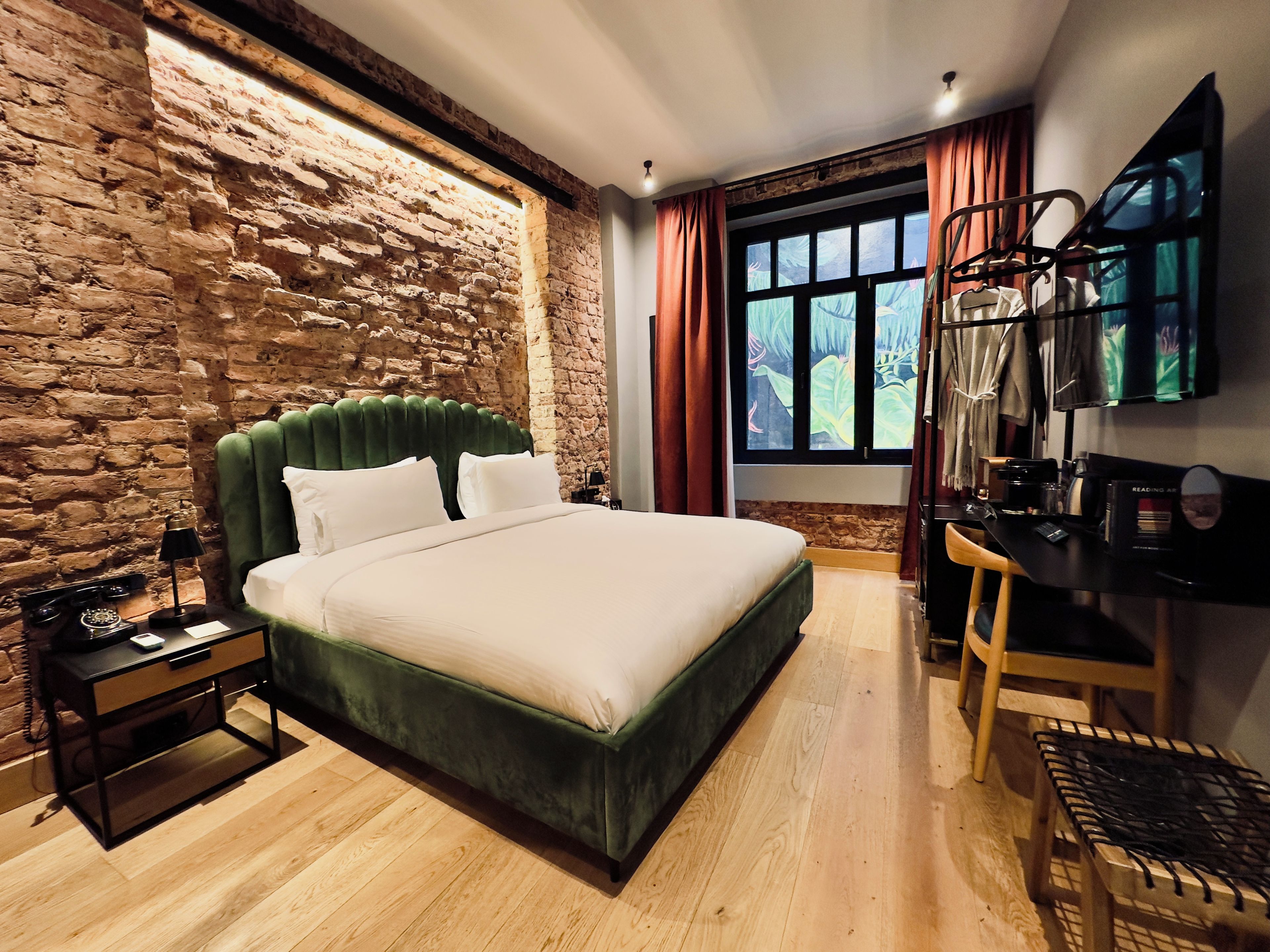 A bedroom with a brick wall, a green upholstered bed, a wooden floor, a desk with a chair, and a window with curtains.