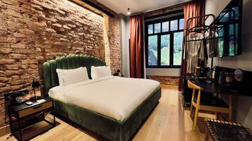 A bedroom with a brick wall, a green upholstered bed, a wooden floor, a desk with a chair, and a window with curtains.