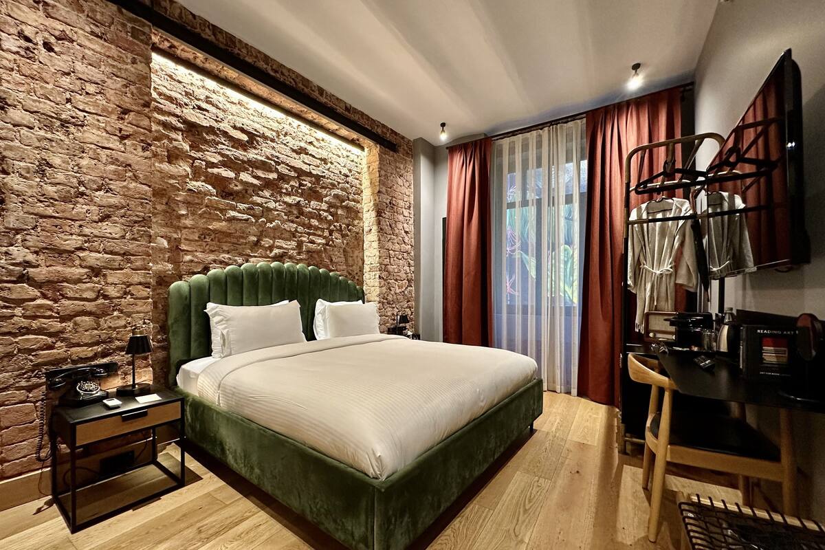 Quarto com parede de pedra, cama estofada em verde, escrivaninha de madeira e uma cadeira.