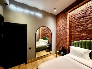Quarto moderno com parede de tijolos, cabeceira estofada em verde, piso de madeira e cama com roupa de cama branca.