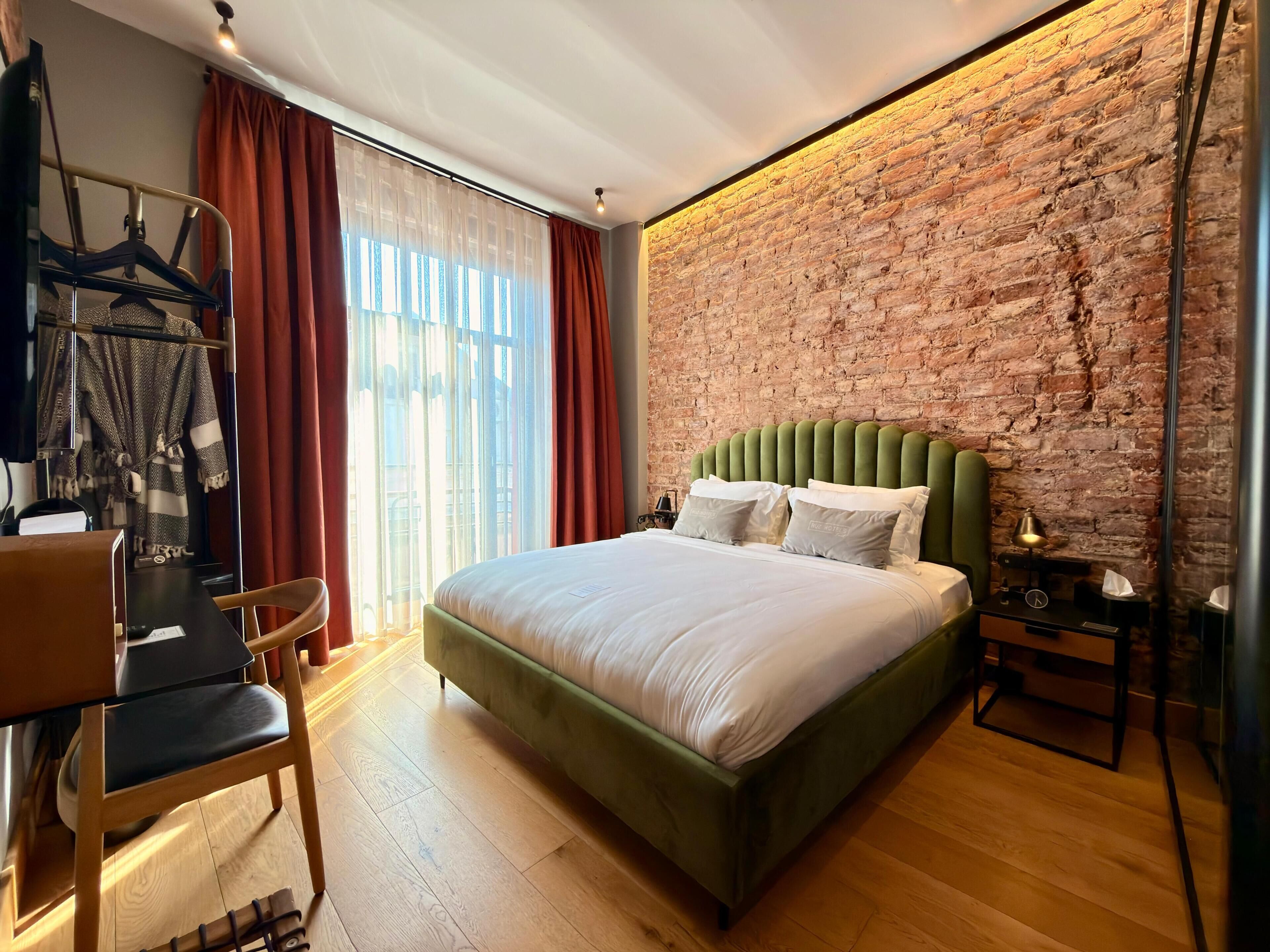 A hotel room with a brick wall, a bed with green upholstered headboard, a wooden floor, a desk with a chair, and a window with curtains.