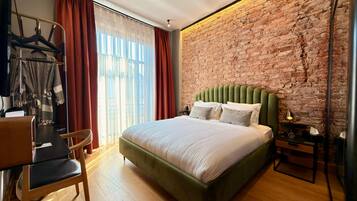 A hotel room with a brick wall, a bed with green upholstered headboard, a wooden floor, a desk with a chair, and a window with curtains.