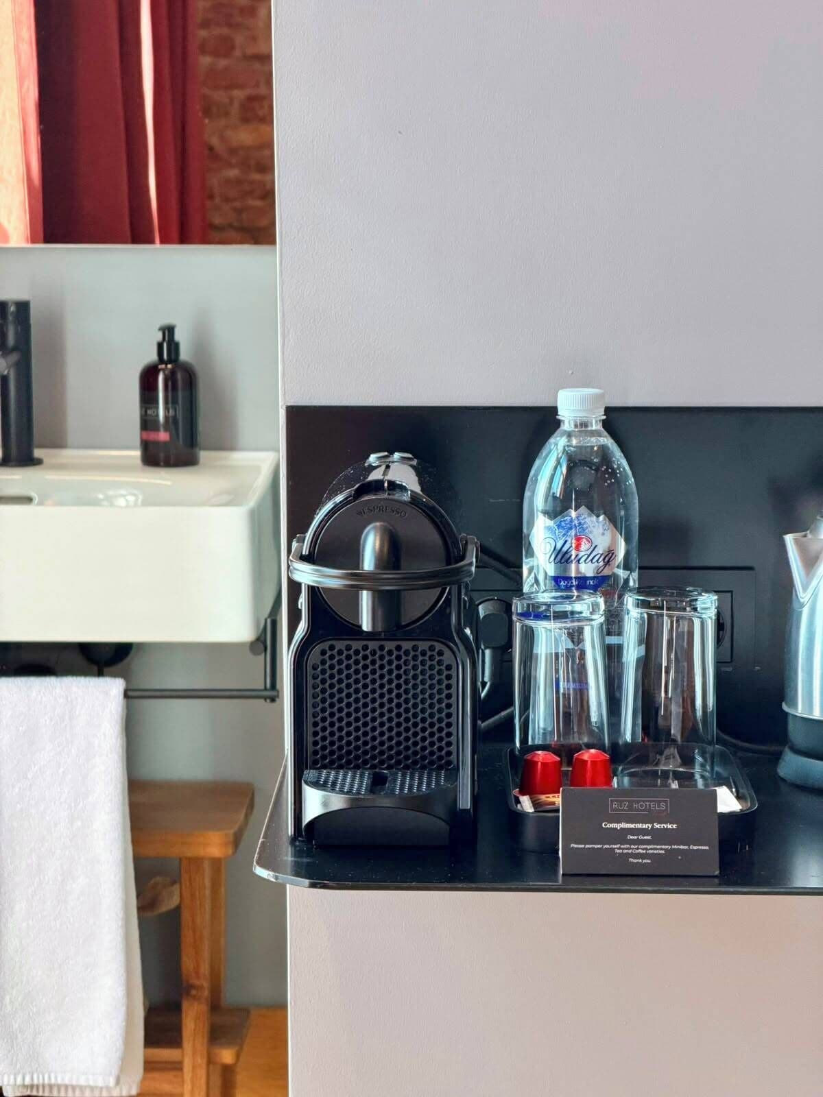 A coffee machine, a water bottle, and a glass carafe on a counter with a towel and a wooden stool nearby.