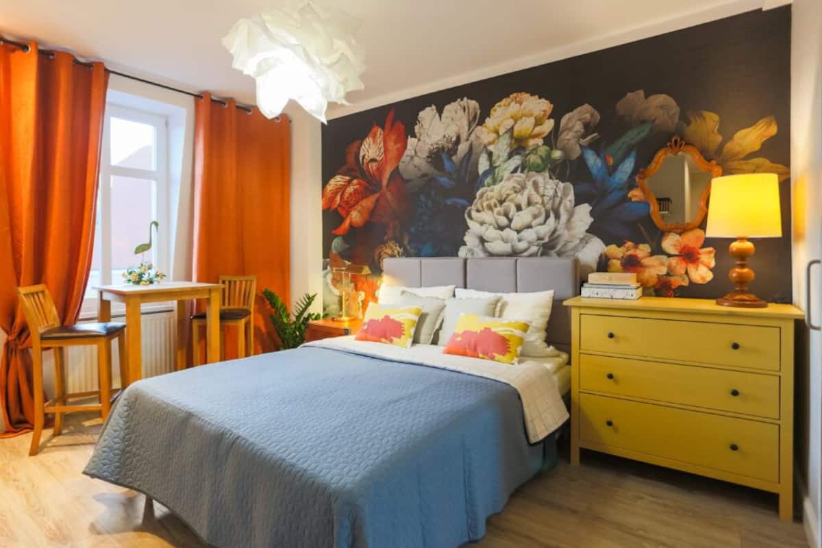 A bedroom with a bed, a desk, a chair, a dresser, and a large floral mural on the wall.