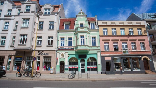 Une rangée de bâtiments historiques aux styles architecturaux distincts, dont un bâtiment à la façade verdoyante ornée de fenêtres en arc de cercle et un bâtiment rose doté d’un balcon.