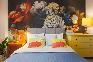 A bedroom with a large floral mural on the wall, a bed with patterned pillows, a bedside table with a lamp, and a nightstand with a mirror.