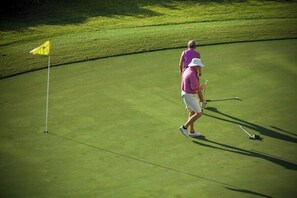 A golf course with a flagstick and a golfer in a purple shirt and white hat.