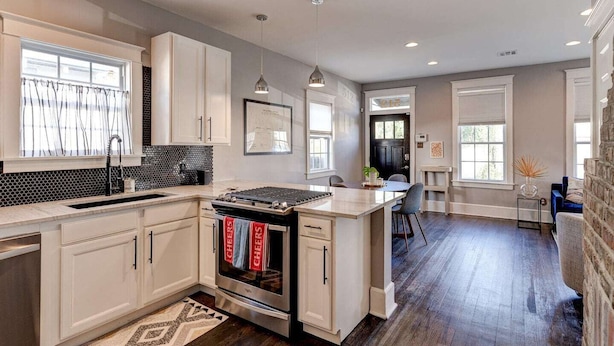 A modern kitchen with white cabinetry, a stainless steel stove, and a dining area with a blue chair. - Charming 3BR Uptown home with fenced yard. Walk to Magazine St, near streetcar & FQ. (New Orleans)
