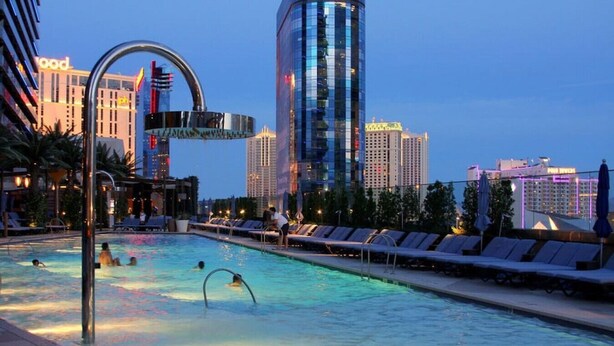 A swimming pool with lounge chairs and a water slide, surrounded by high-rise buildings and palm trees. - ON THE STRIP! | Classic Comfort - 2 Bedroom Condo (Las Vegas)