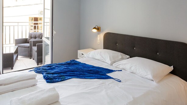 Hotelkamer met een bed, witte kussens, een blauwe deken, een nachtkastje, een wandlamp en uitzicht op een balkon met rieten meubels.