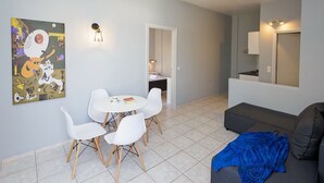 Een moderne woonkamer met een grijze bank, een blauwe deken en een witte eettafel met vier stoelen. Er hangt een schilderij aan de muur, en een deur leidt naar een badkamer.
