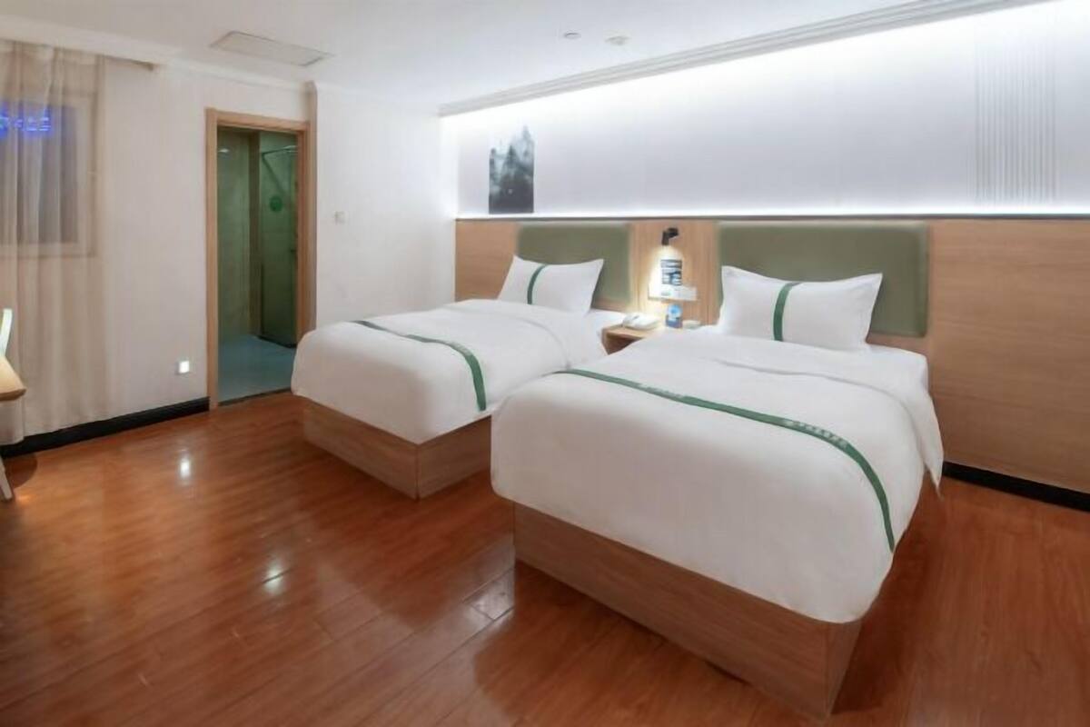 A hotel room with two beds, a wooden floor, a nightstand, a lamp, and a door leading to a bathroom.