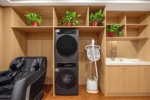 A compact room with a black massage chair, a stacked washer and dryer, a small sink, and shelves with potted plants. - GreenTree Inn (Hefei Datang International Honggang Metro Station) (Hefei)