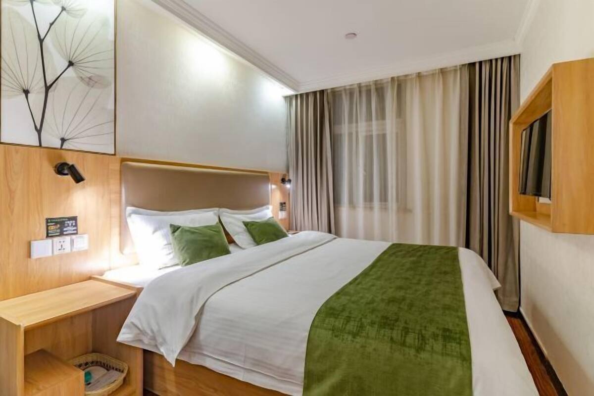 A hotel room with a bed, a wooden nightstand, a wall-mounted TV, and a window with curtains.
