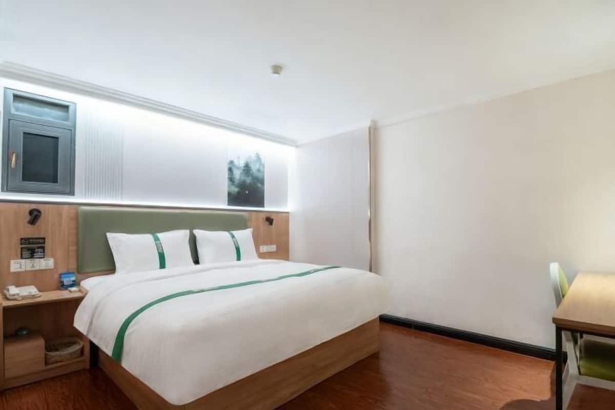 A hotel room with a bed, a desk, a nightstand, a window, and a wall-mounted TV.