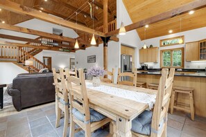 A spacious living area with a wooden dining table, chairs, a sofa, and a kitchen with wooden cabinets and a stainless steel refrigerator.