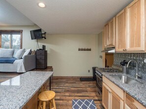 A compact kitchen with granite countertops, wooden cabinets, a built-in stove, and a TV mounted on the wall. - Brookside II A-215 - Sunday River Condo (Newry)