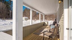 A covered porch with a view of a snowy landscape, wooden flooring, and a door to the right. - Sunday River Getaway (Newry)