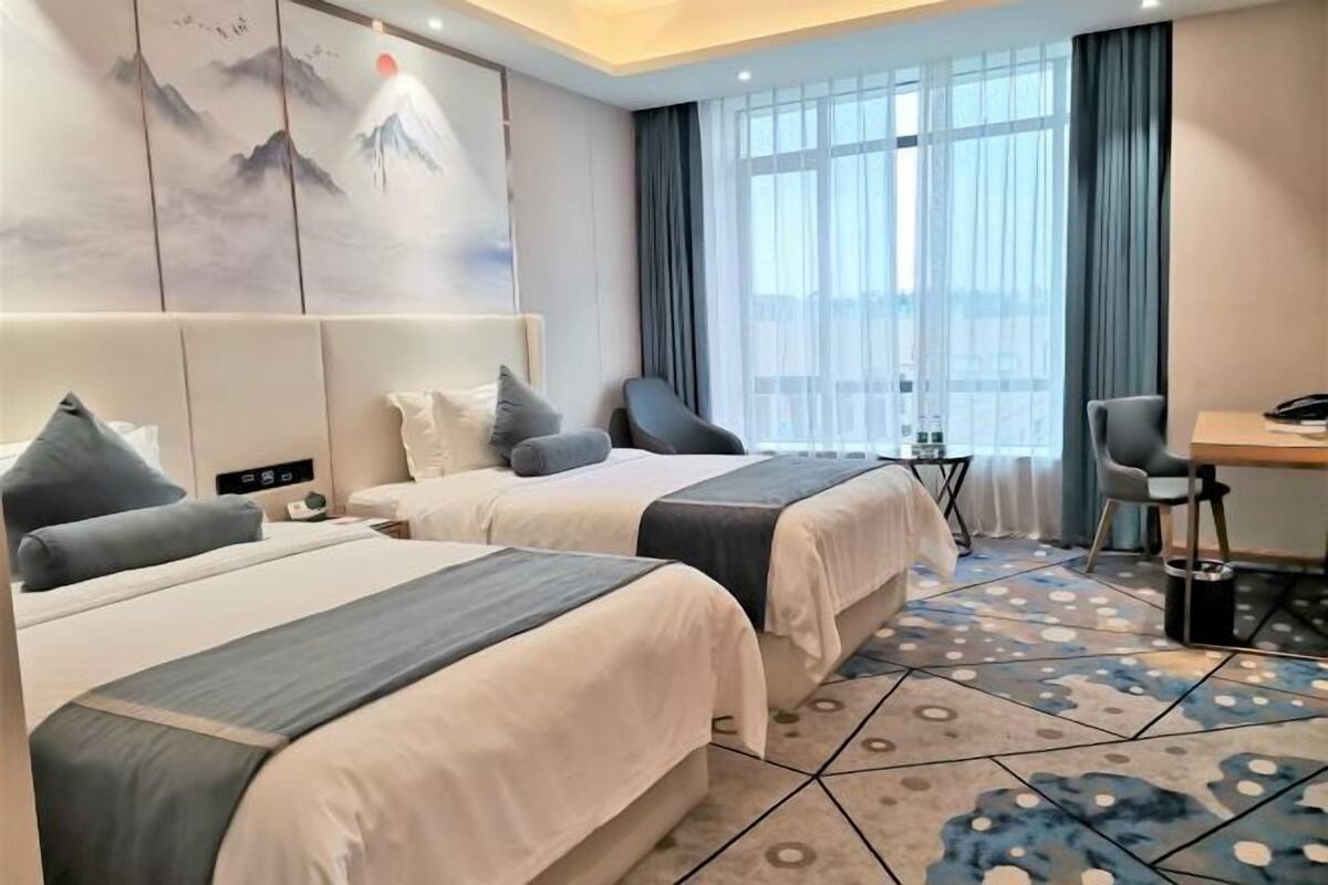 A hotel room with two beds, a desk, a chair, a small table, and a large window with curtains.