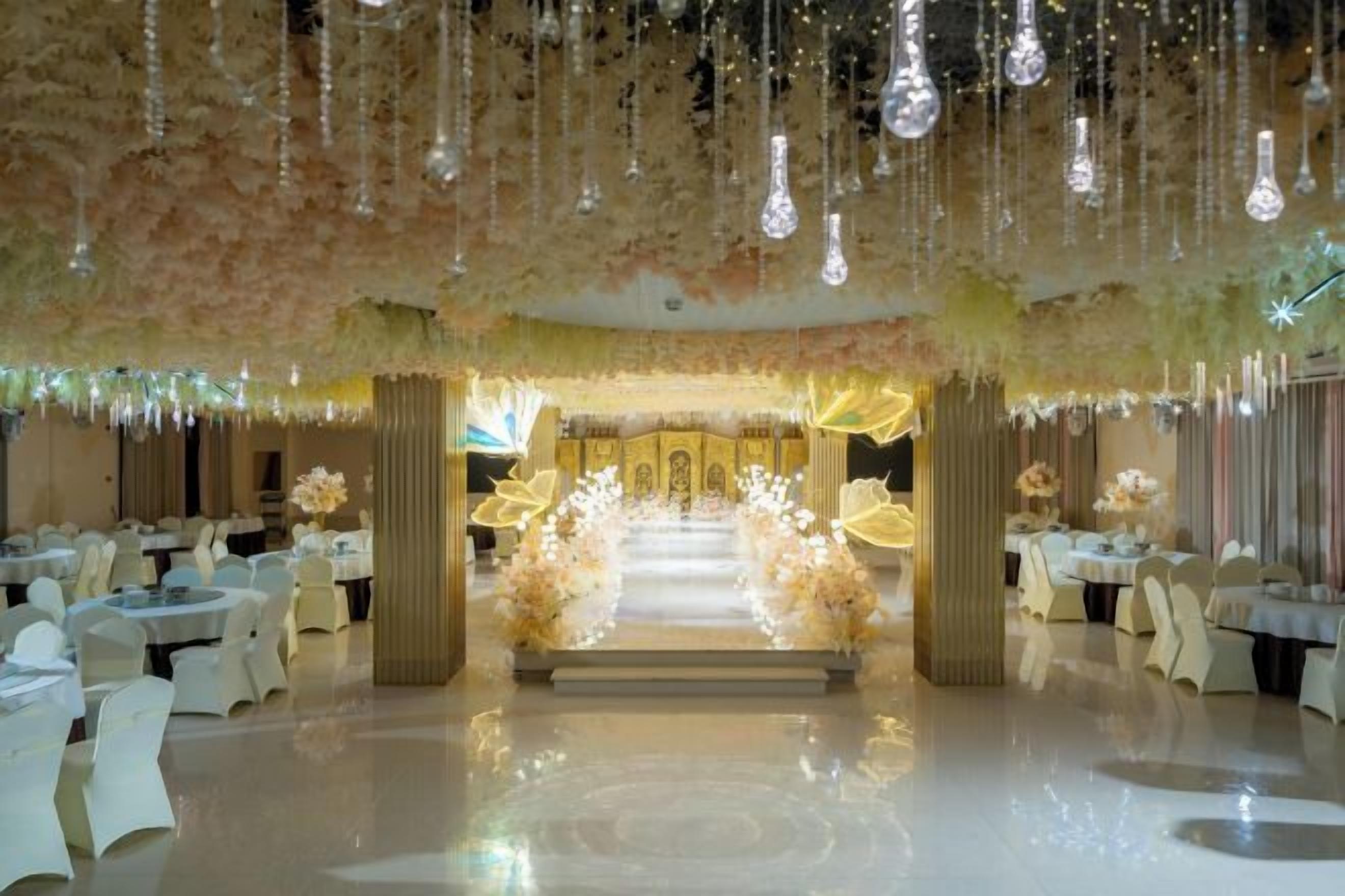 Un salón de banquetes con mesas dispuestas para un evento formal, un pasillo central que conduce a un escenario decorado y lámparas colgantes.