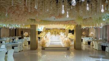 Un salón de banquetes con mesas dispuestas para un evento formal, un pasillo central que conduce a un escenario decorado y lámparas colgantes.