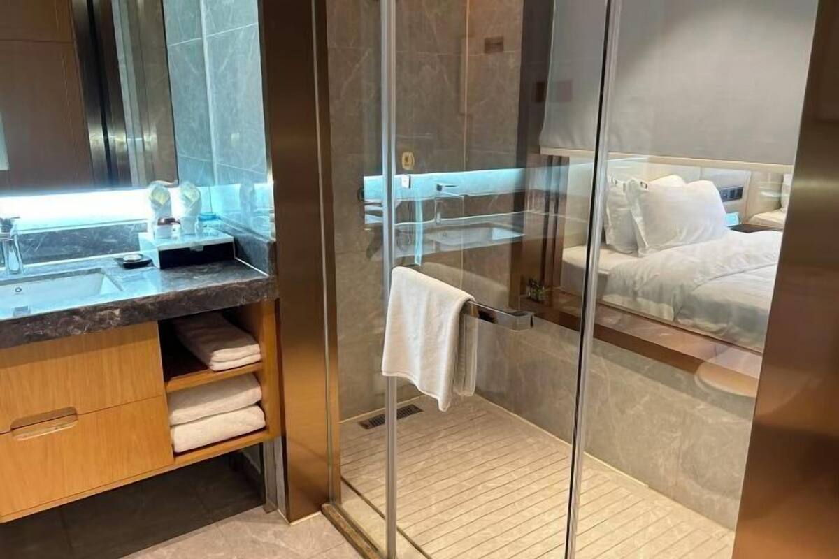 A modern bathroom with a glass shower enclosure, a wooden vanity, and a bed visible through the glass.