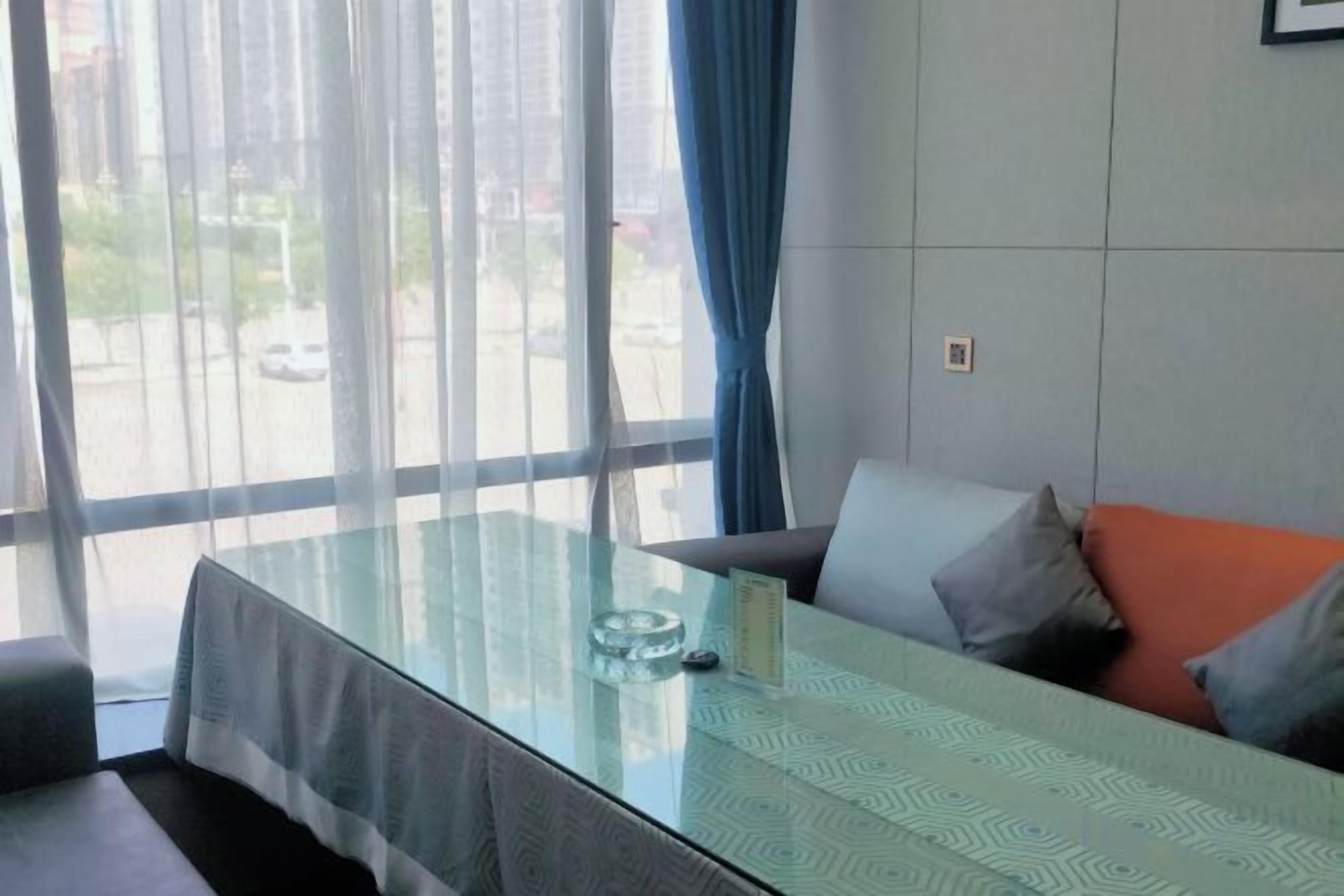 A modern room with a glass table, a sofa with cushions, and a view of the street through large windows.
