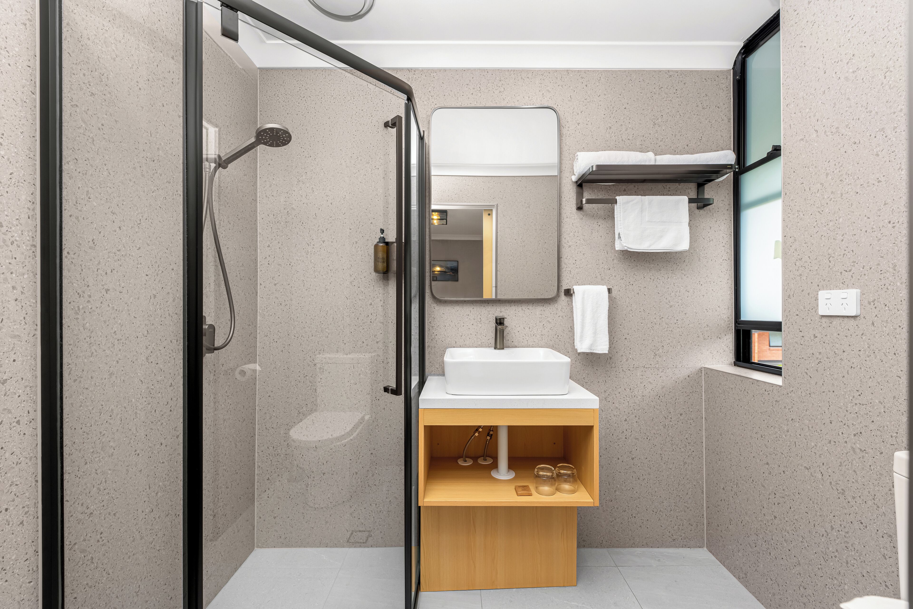 Un baño moderno con ducha envuelta en vidrio, un lavamanos blanco y un estante de madera.