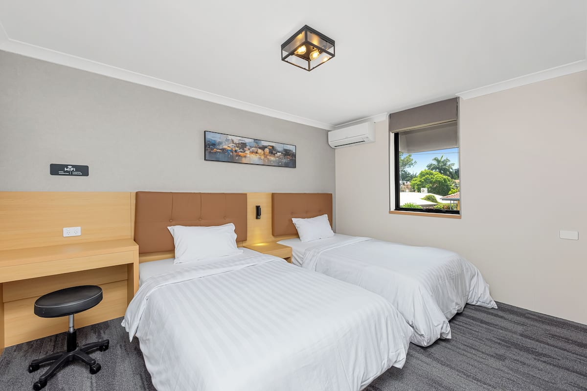 A hotel room with two beds, a desk, a chair, and a window with a view of greenery.