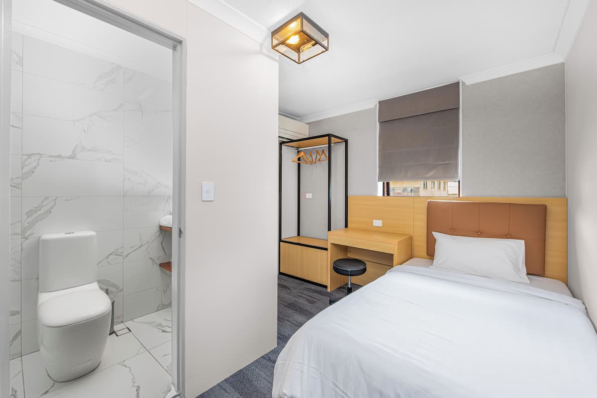 A hotel room with a toilet, a bed, a wooden nightstand, a mirror, and a ceiling light fixture.