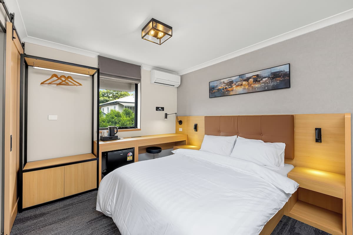 A modern hotel room with a large bed, a desk with a computer, a small kitchenette, and a window with a view of greenery.