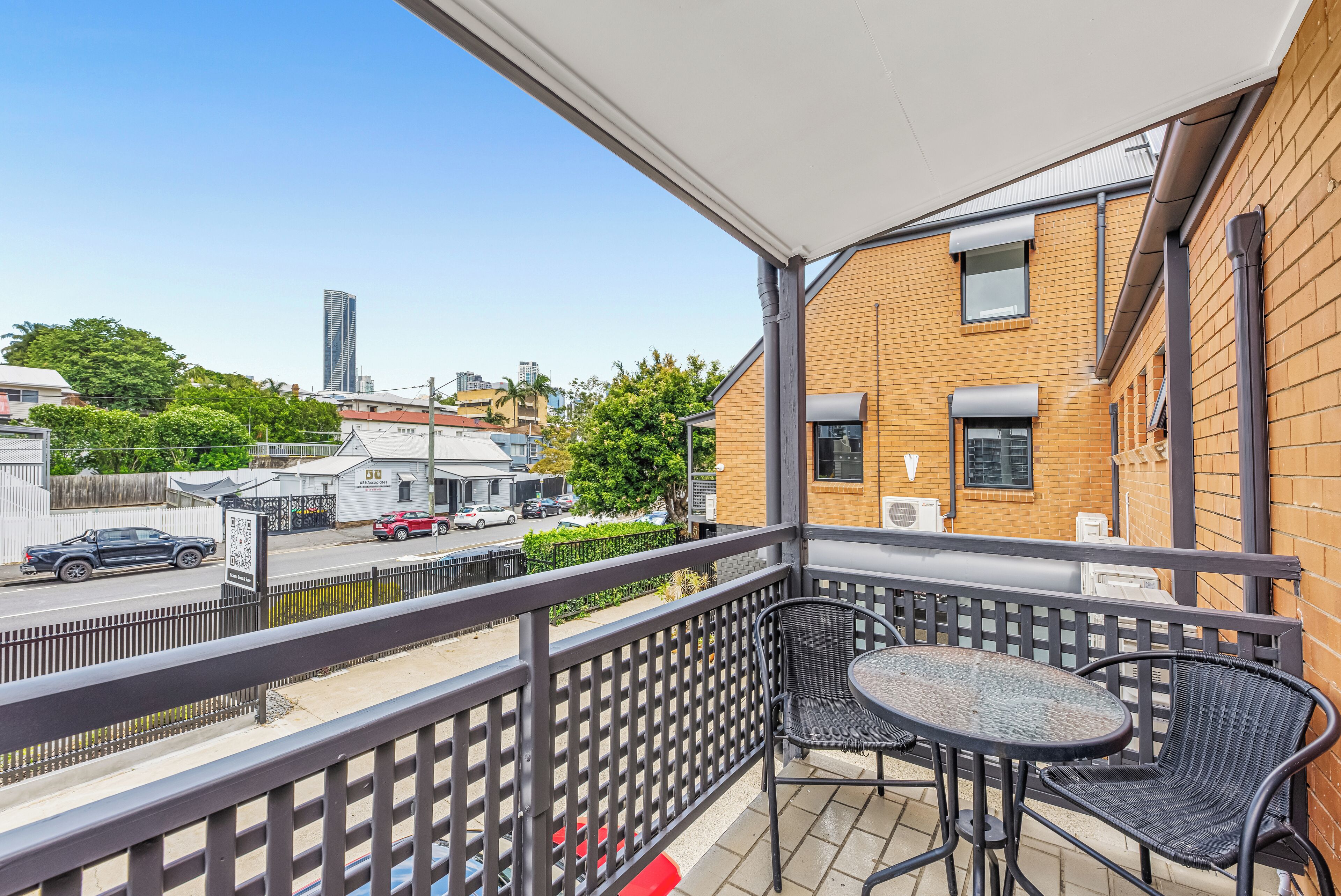 Varanda com duas cadeiras e uma mesa, com vista para uma rua com carros e edifícios.