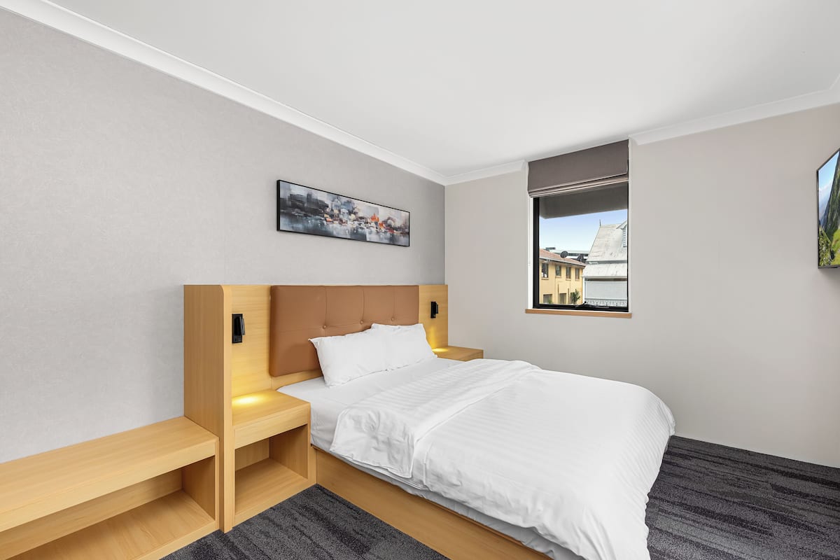 A modern hotel room with a bed, wooden nightstands, a painting, a window with a view, and a small table.