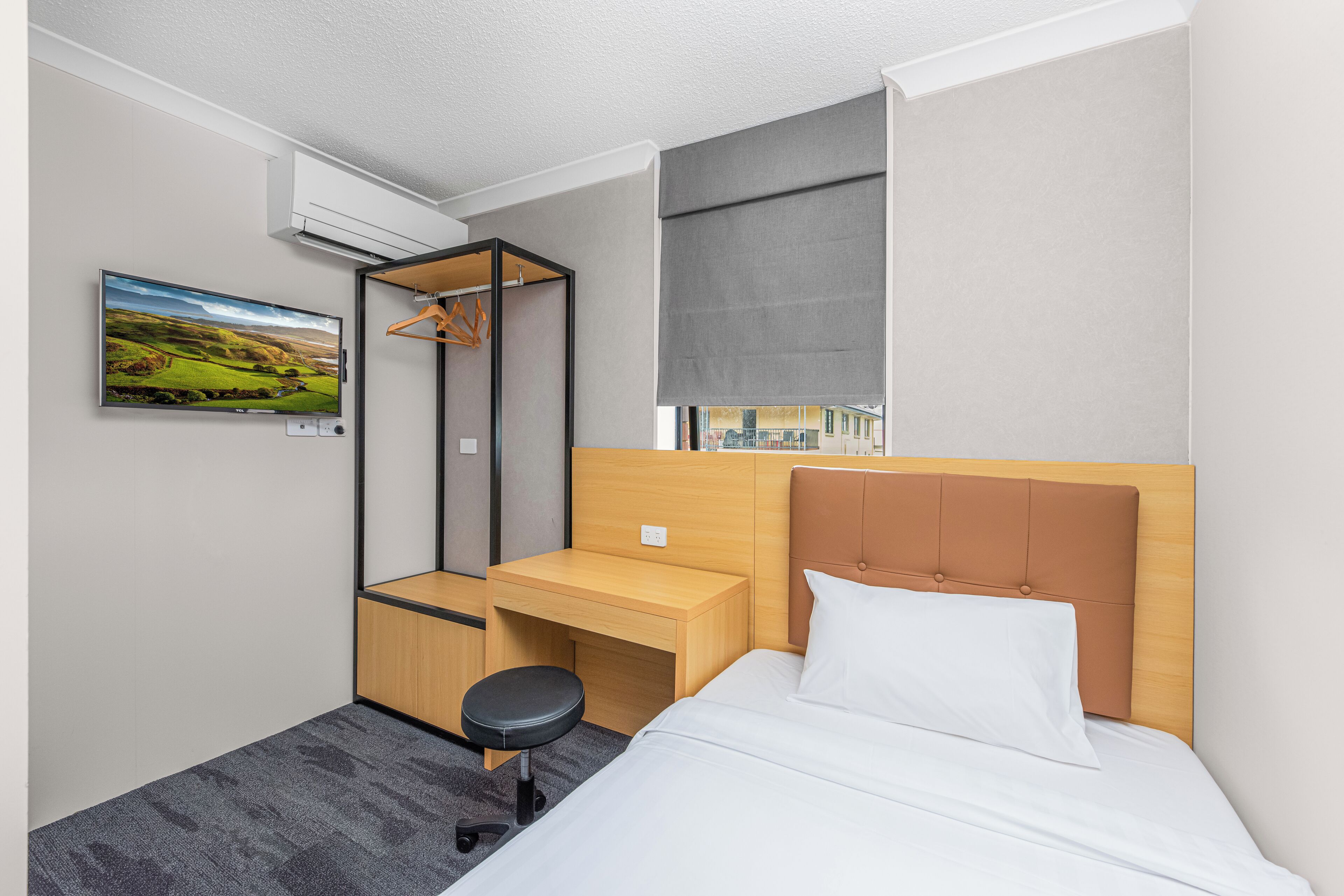 Moderni hotellihuone, jossa on sänky, työpöytä, tuoli, seinälle kiinnitetty naulakko ja suuri ikkuna, josta on näkymä rakennuksiin.