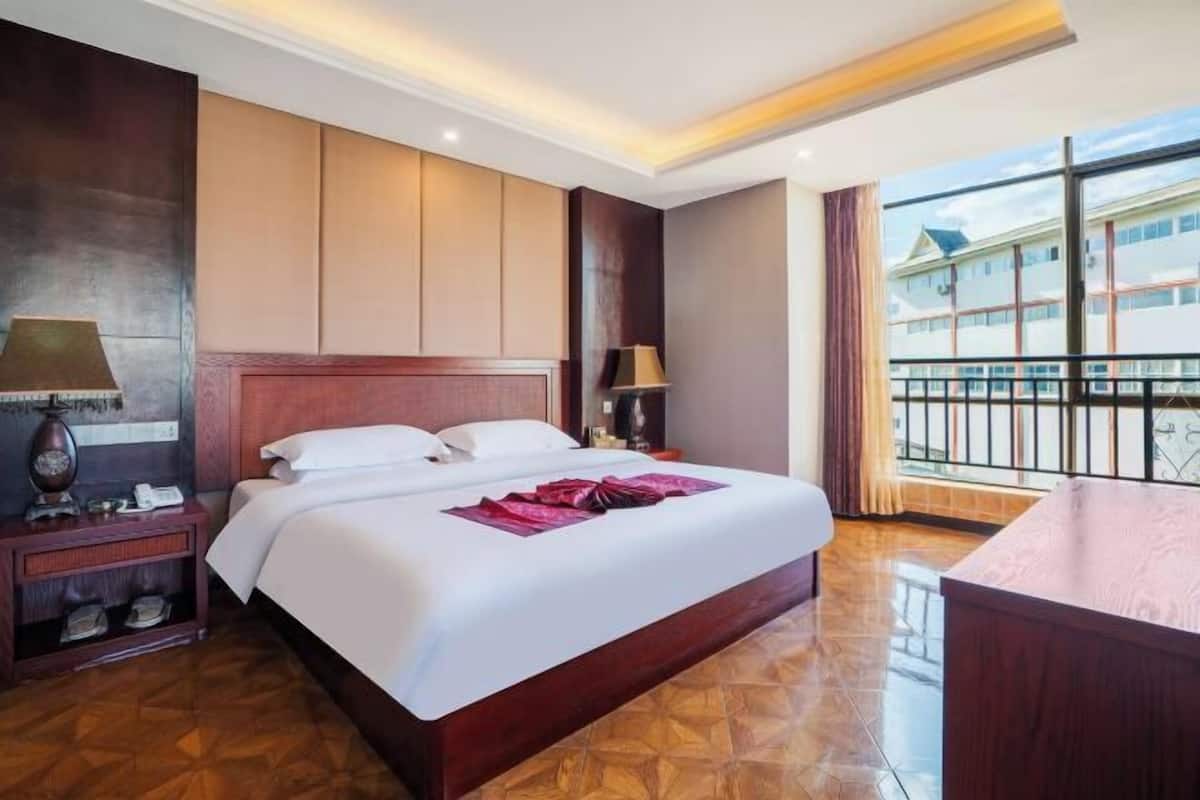 Hotelkamer met een groot bed, houten hoofdeinde, nachtkastje met lamp en uitzicht op gebouwen vanaf een balkon.
