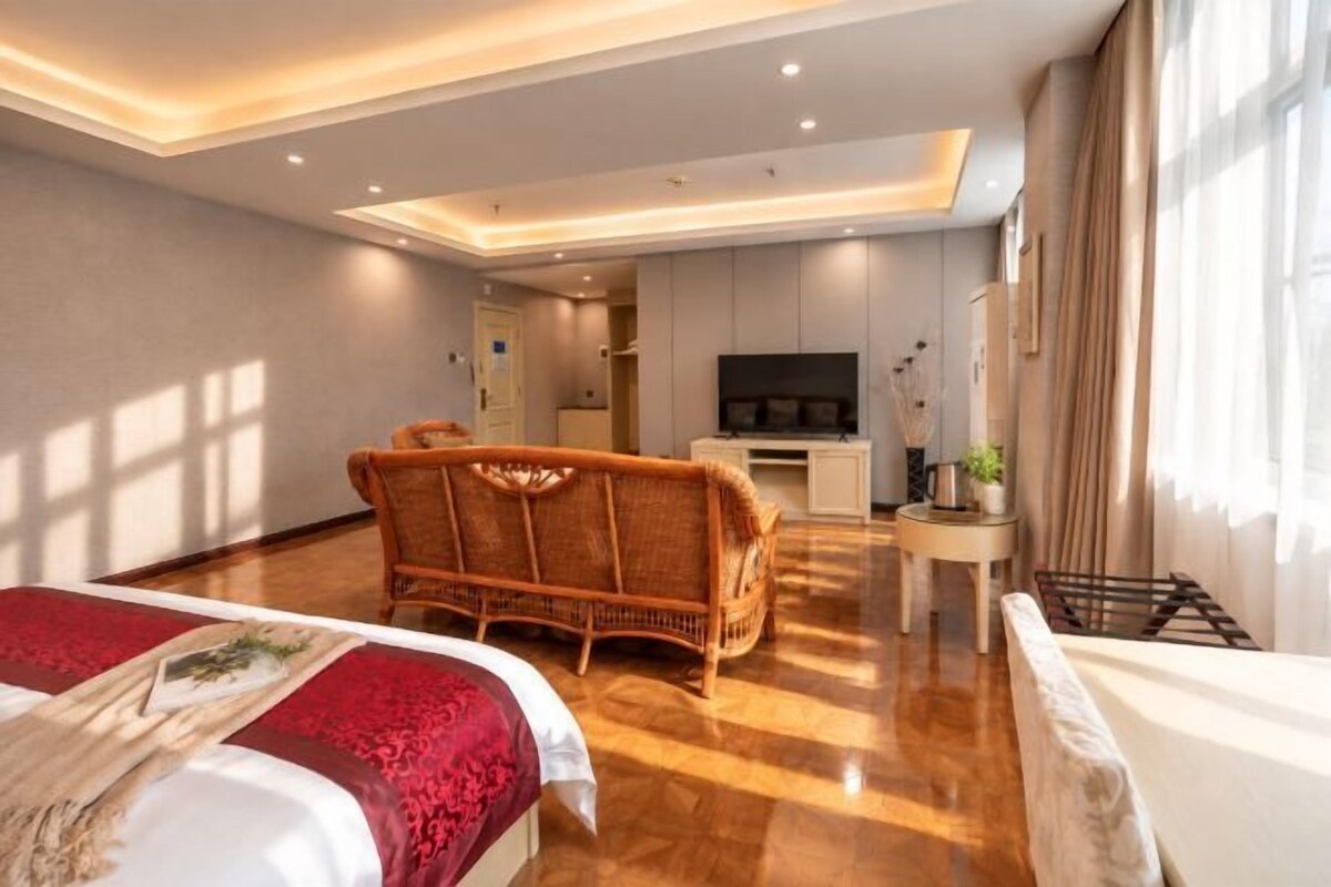 A hotel room with a bed, a wicker chair, a small table, a TV, and a kitchenette in the background.