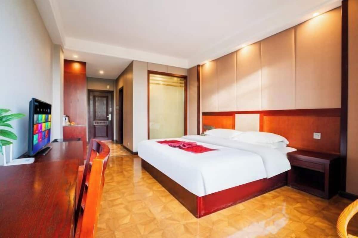 Een hotelkamer met een houten vloer, een bed met wit beddengoed en een rode accentkleur, een nachtkastje en een tv op een houten plank.