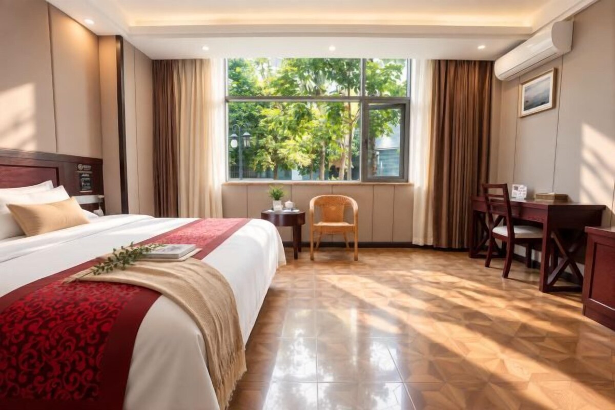 A hotel room with a large bed, a desk, and a chair. There is a window with a view of trees.