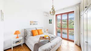 Un dormitorio con una cama, una mesita de noche con lámpara, una bandeja con desayuno y un amplio ventanal con vistas a zonas verdes.