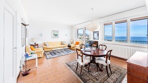 Una sala de estar luminosa con sofá, mesa de centro y mesa de comedor. Amplias ventanas ofrecen vistas al mar.