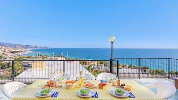 Tavolo imbandito con piatti, bicchieri e cibo, su un balcone con vista sulla costa.