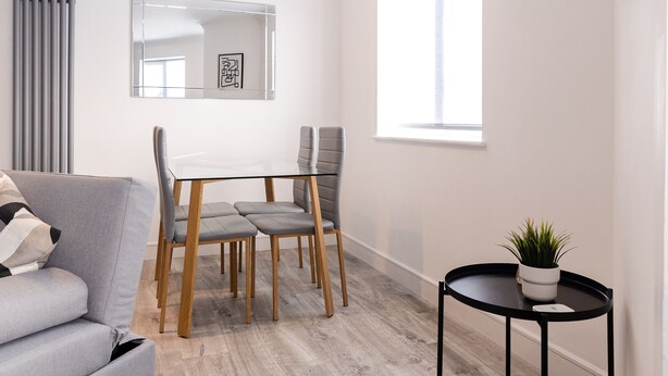 A modern living room with a grey sofa, a dining table with chairs, a small black side table with a plant, and a large window providing natural light. - Luxury, Modern Apartment w/ Netflix in Zone 2 (London)